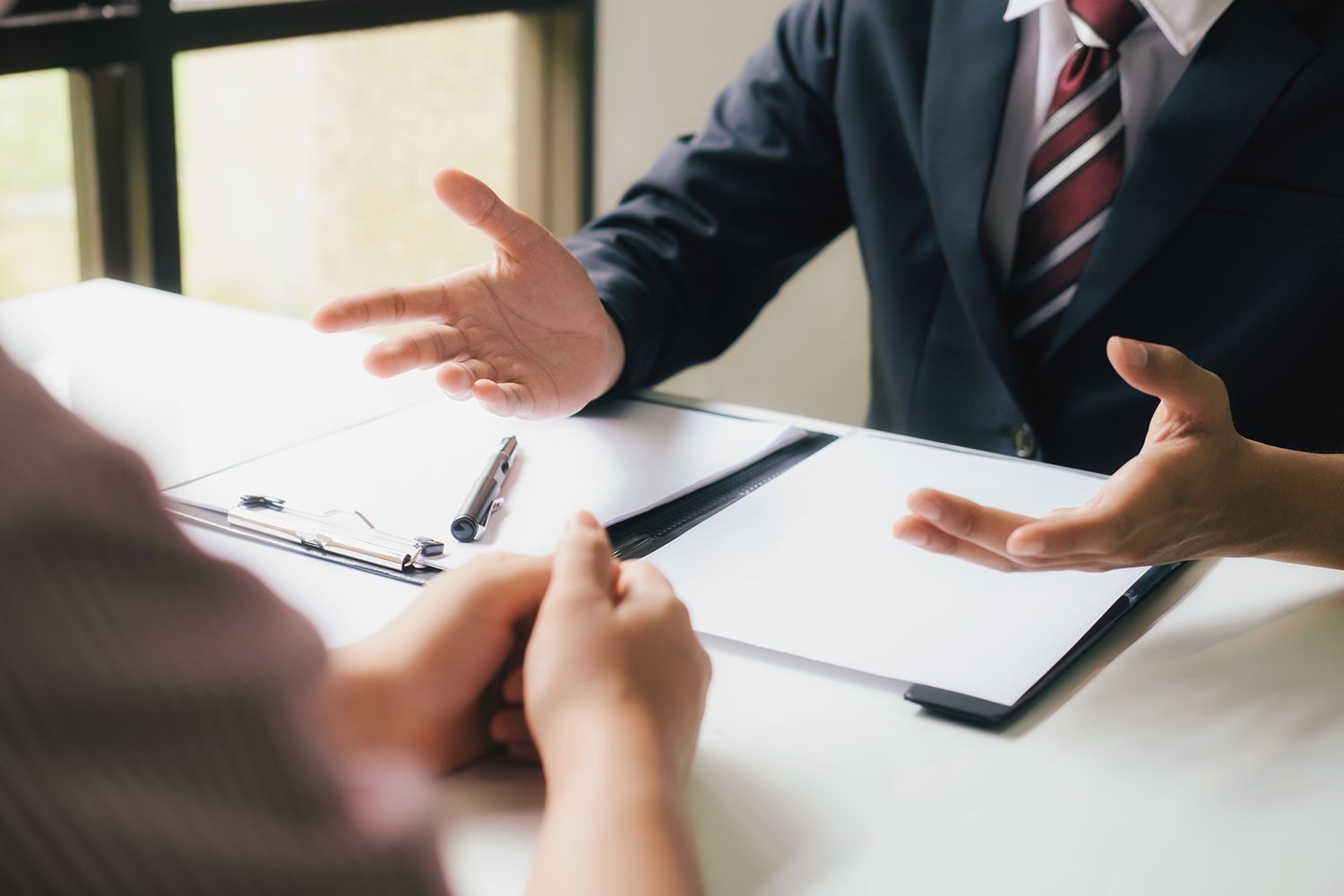 Two people at a desk; one in a suit gestures, the other listens and has hands clasped. Documents and pen visible. Two people at a desk; one in a suit gestures, the other listens and has hands clasped. Documents and pen visible.