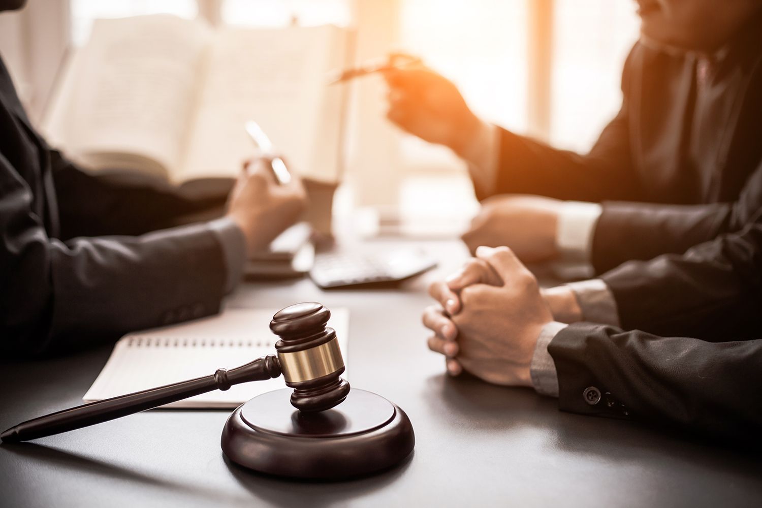 Gavel on a table with lawyers discussing; open book and laptop present. Gavel on a table with lawyers discussing; open book and laptop present.