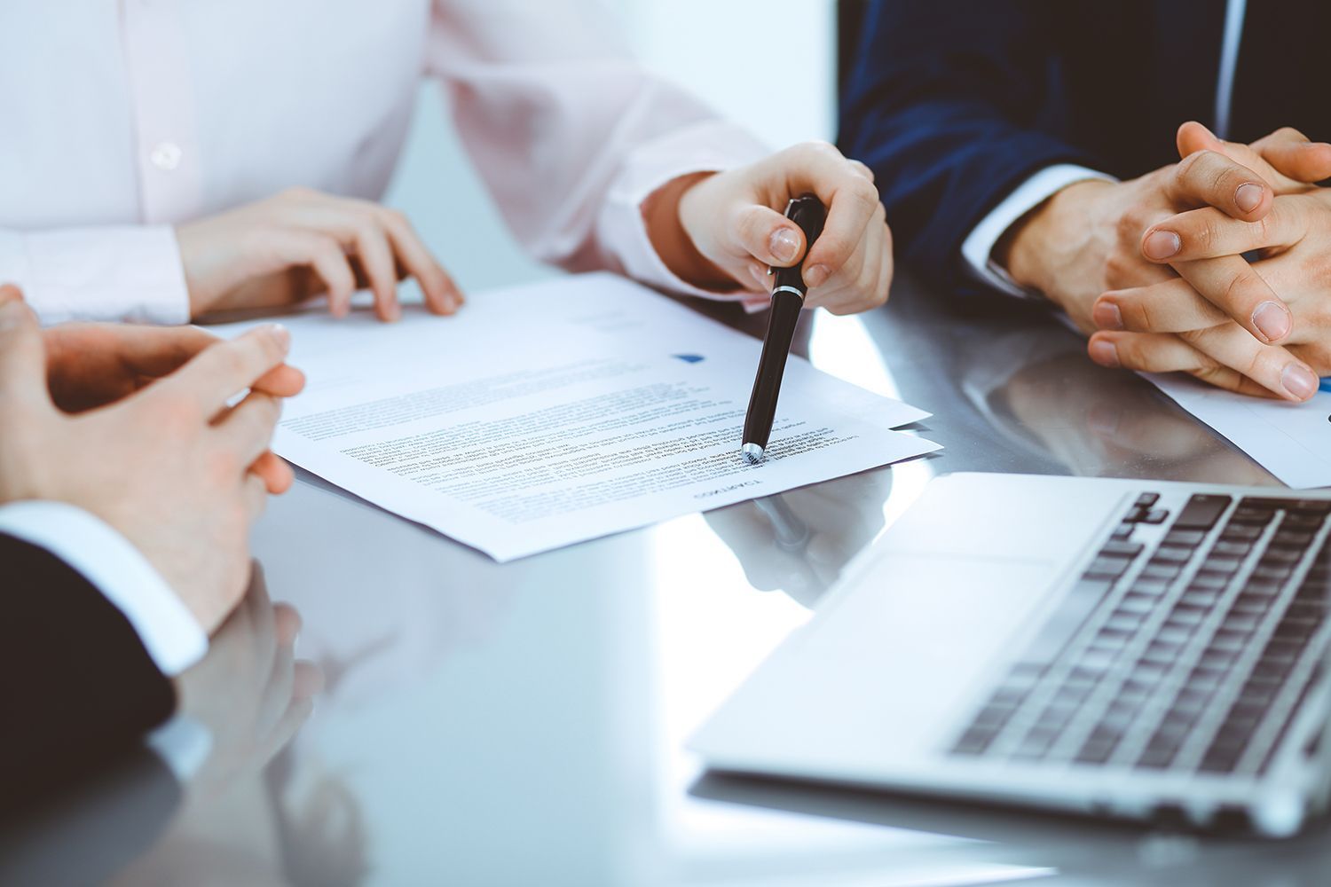 Hands pointing to documents at a table; laptop present, suggesting a business meeting. Hands pointing to documents at a table; laptop present, suggesting a business meeting.