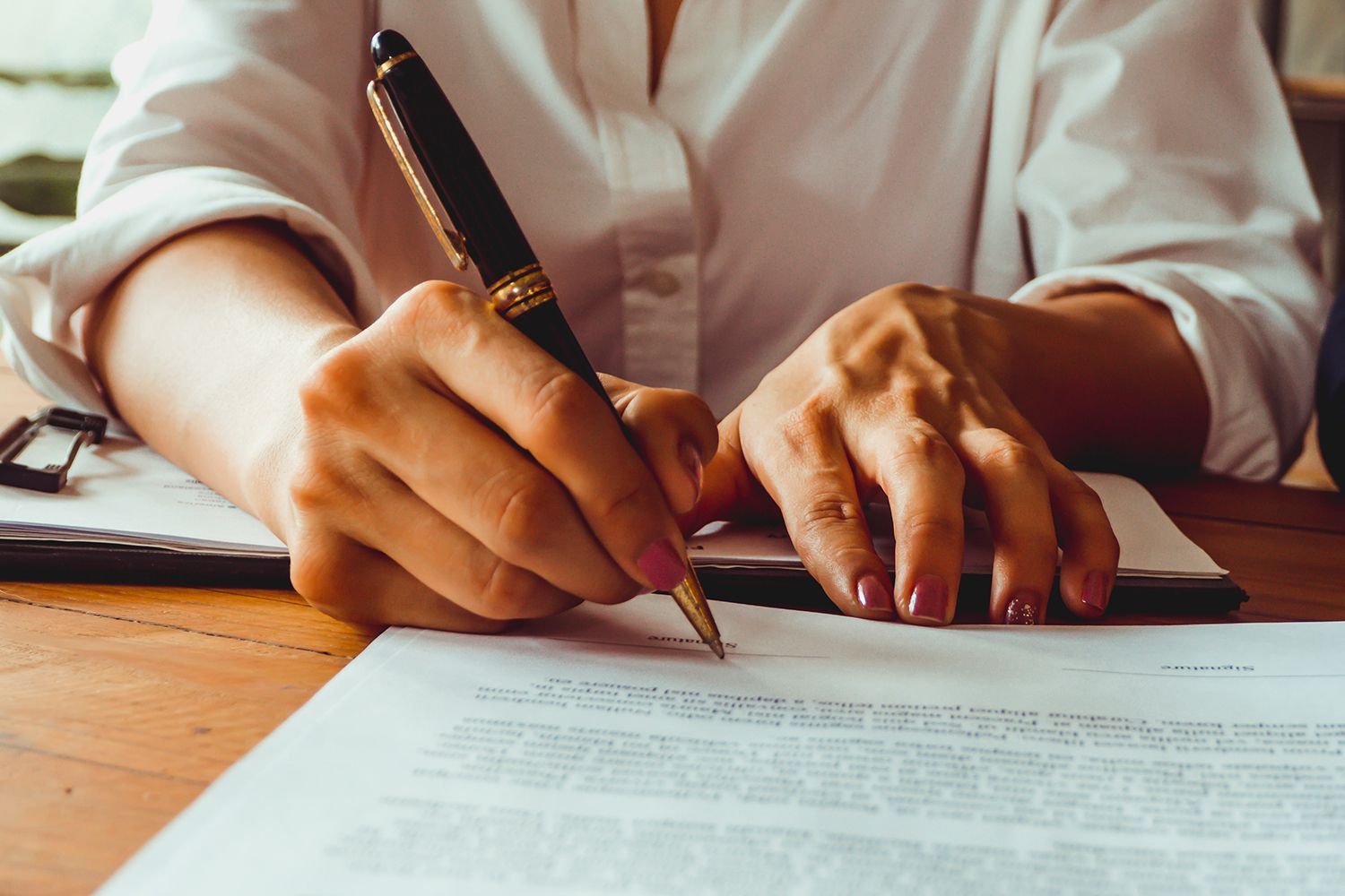 Woman's hands writing with a pen on a document at a wooden table. Woman's hands writing with a pen on a document at a wooden table.
