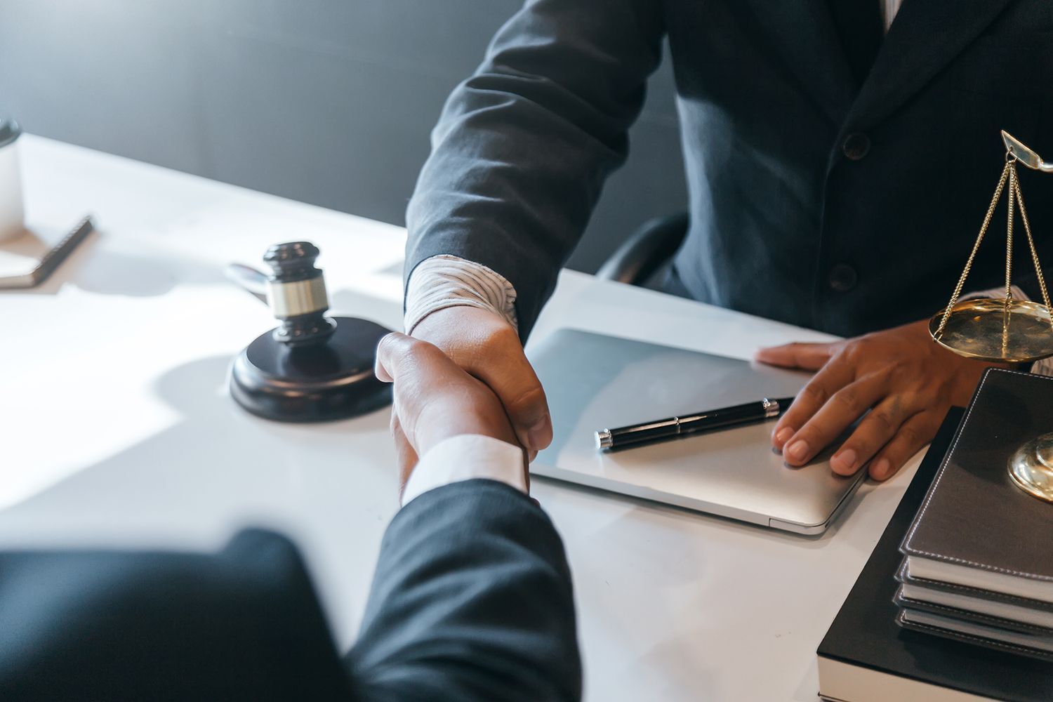 Two people in suits shaking hands over a desk with a gavel and scales of justice.