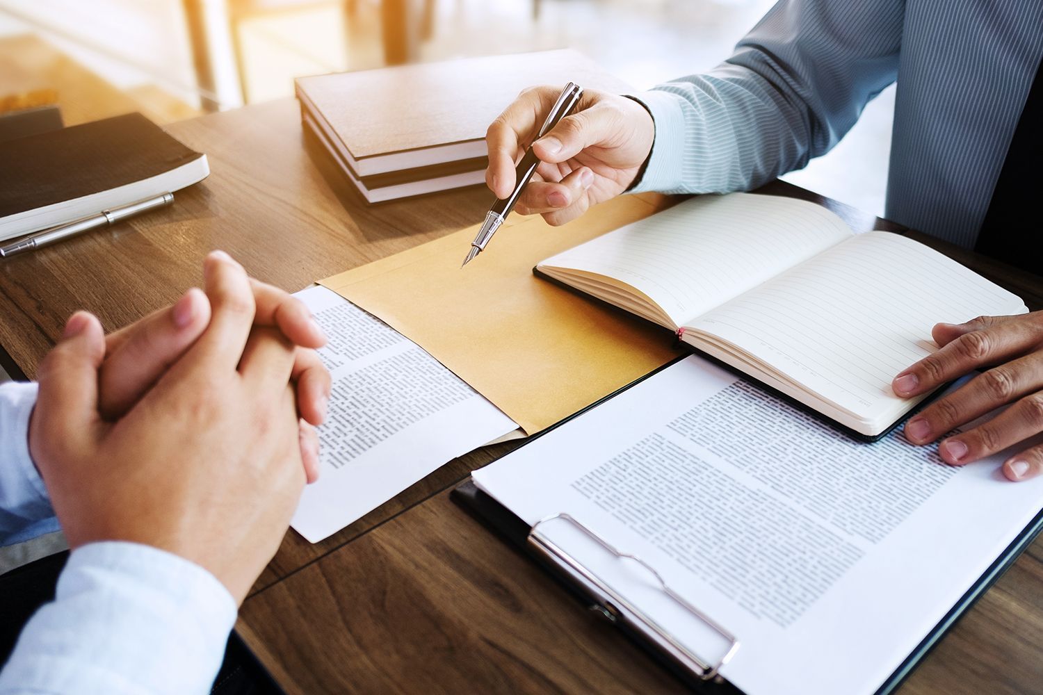 Two people at a desk reviewing documents; one points a pen at a paper while another clasps hands. Two people at a desk reviewing documents; one points a pen at a paper while another clasps hands.