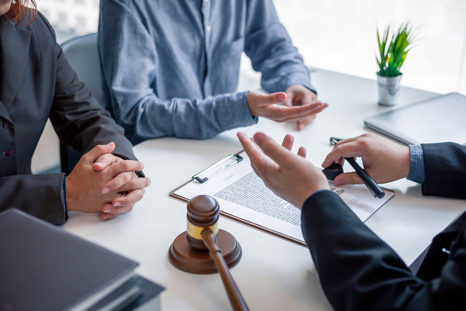 Two people at a desk with a lawyer, discussing documents. Gavel, pen, and a small potted plant on the table. Two people at a desk with a lawyer, discussing documents. Gavel, pen, and a small potted plant on the table.
