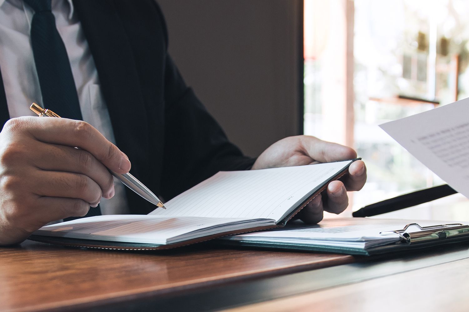 Person in suit writing in notebook with a pen at a desk. Person in suit writing in notebook with a pen at a desk.