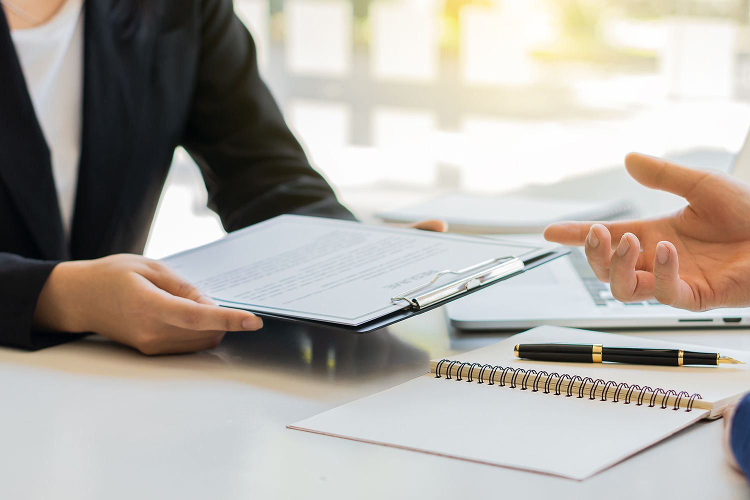 Person in suit handing document to another person, office setting with laptop and notepad.