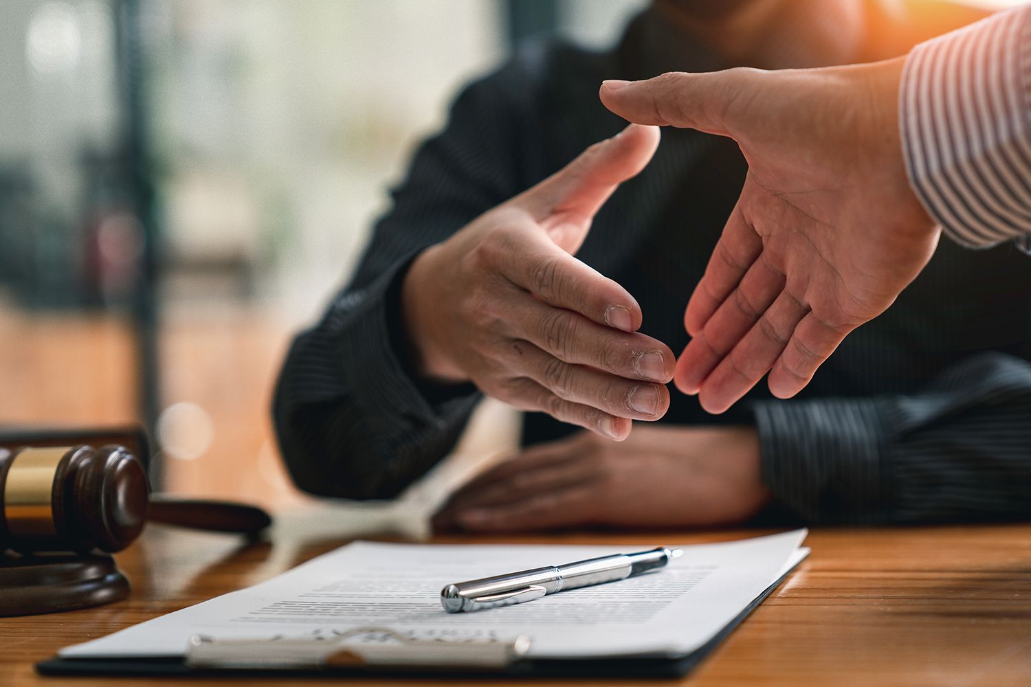 Two hands reaching for a handshake over a contract on a table with a gavel nearby. Two hands reaching for a handshake over a contract on a table with a gavel nearby.