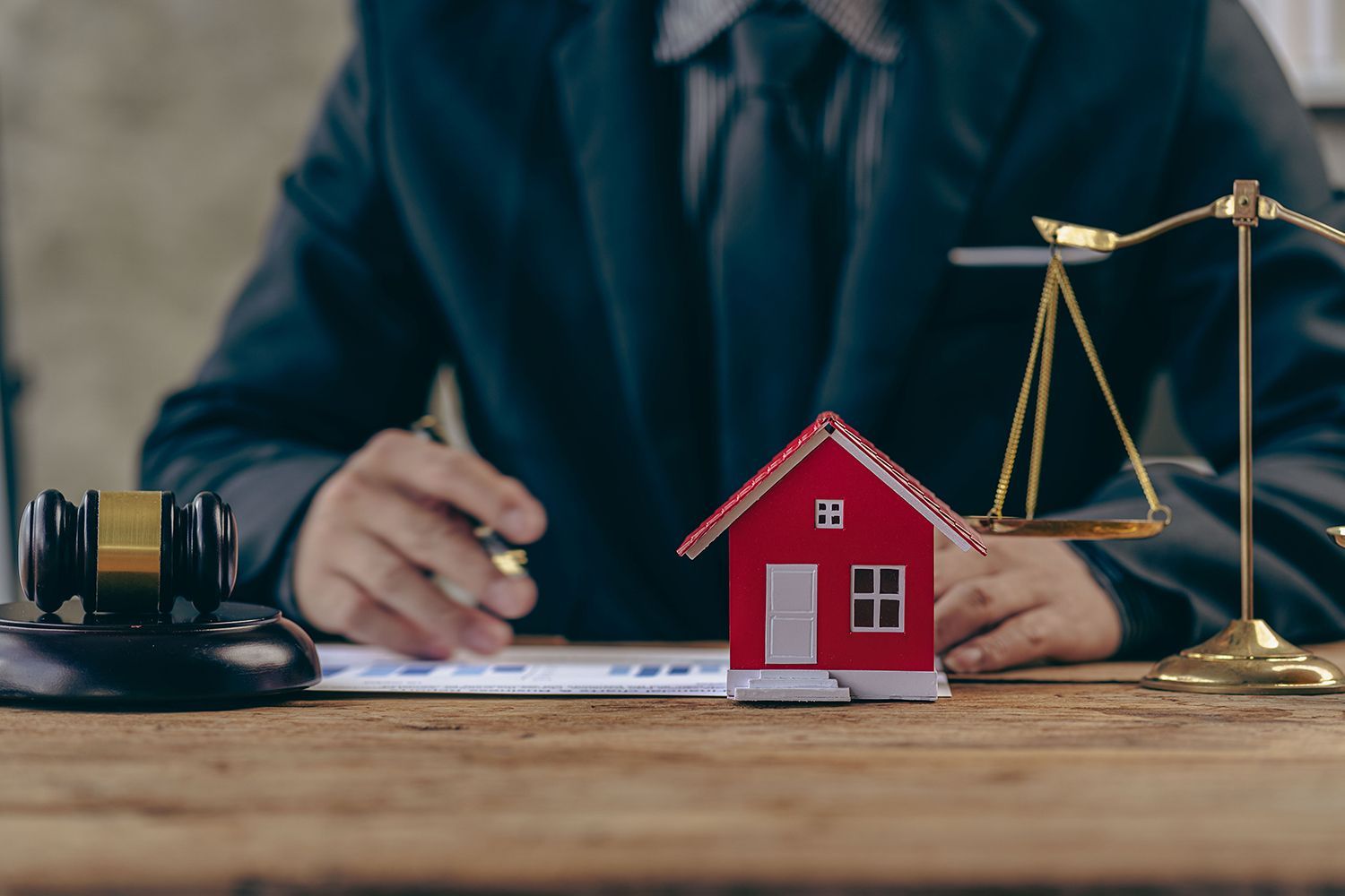 Lawyer at desk with gavel, scales, and red house model; legal documents visible.