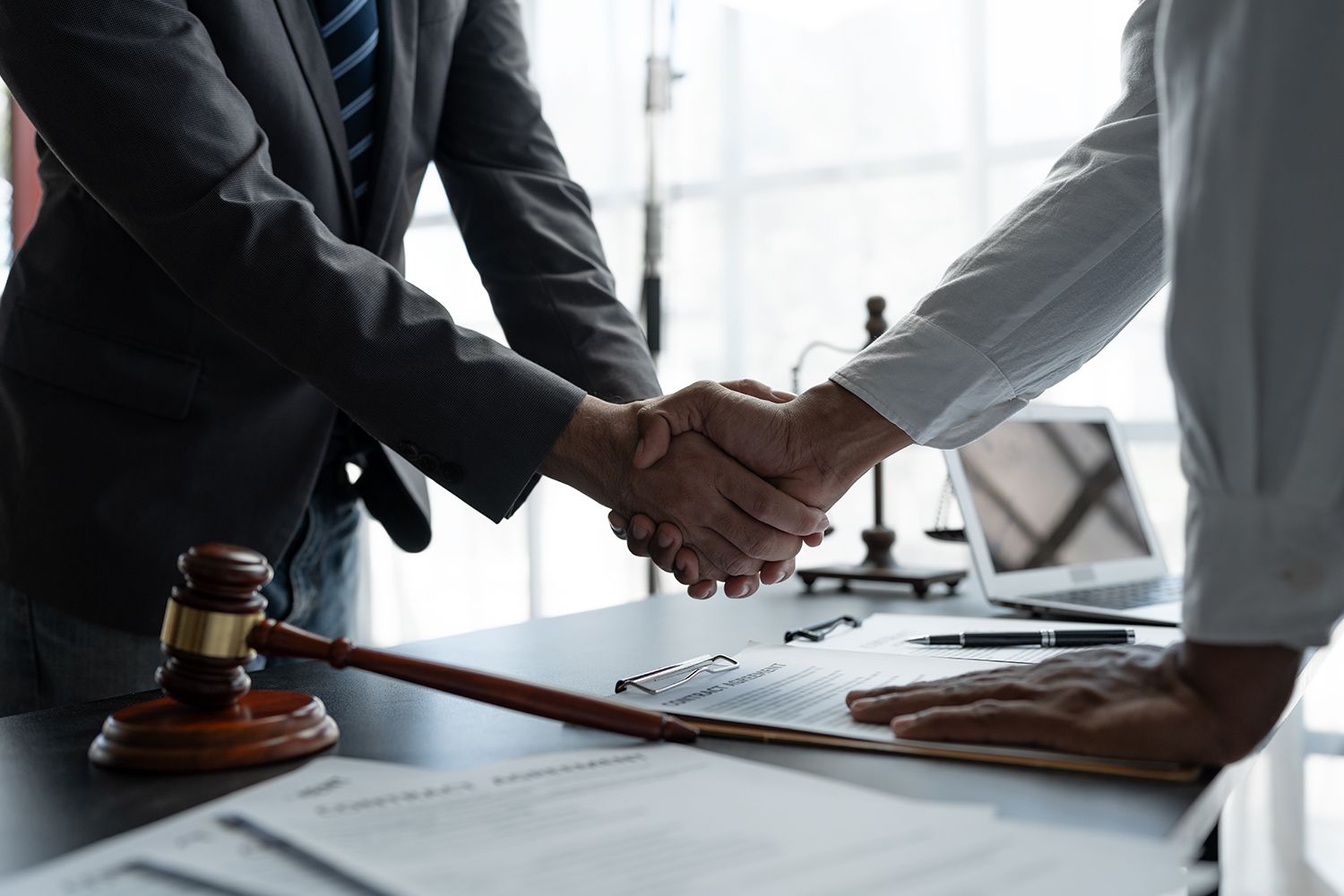 Two people shaking hands over a contract on a desk, gavel and scales of justice in background. Two people shaking hands over a contract on a desk, gavel and scales of justice in background.