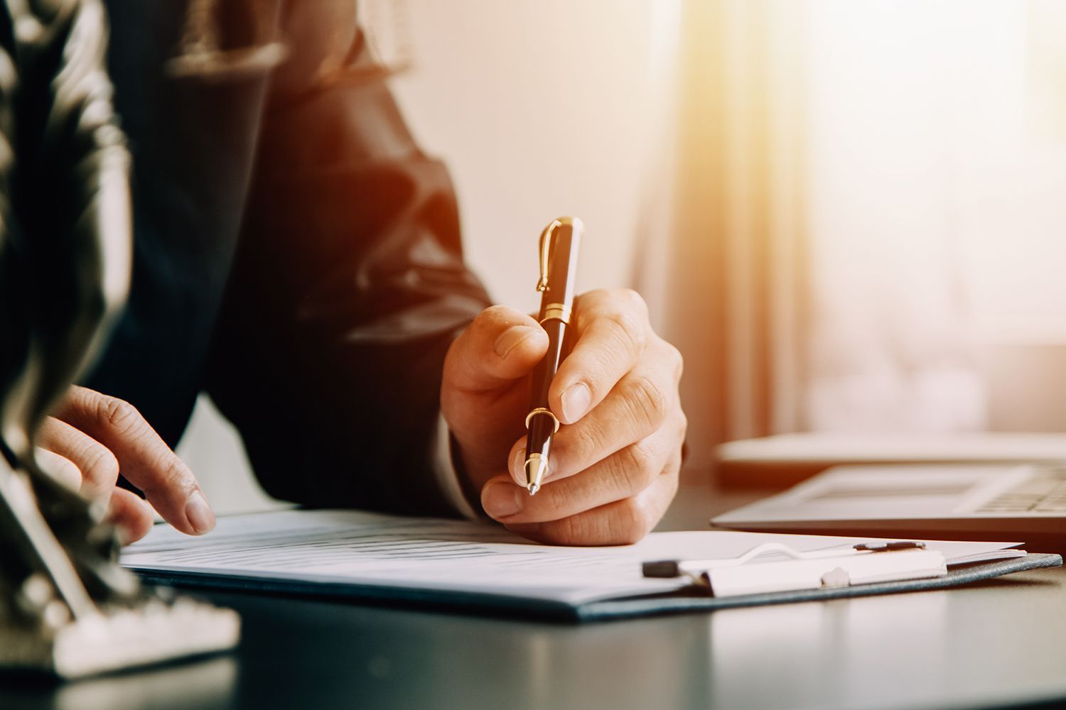 Person in suit signing documents with a pen at a desk, sunlight in background. Person in suit signing documents with a pen at a desk, sunlight in background.
