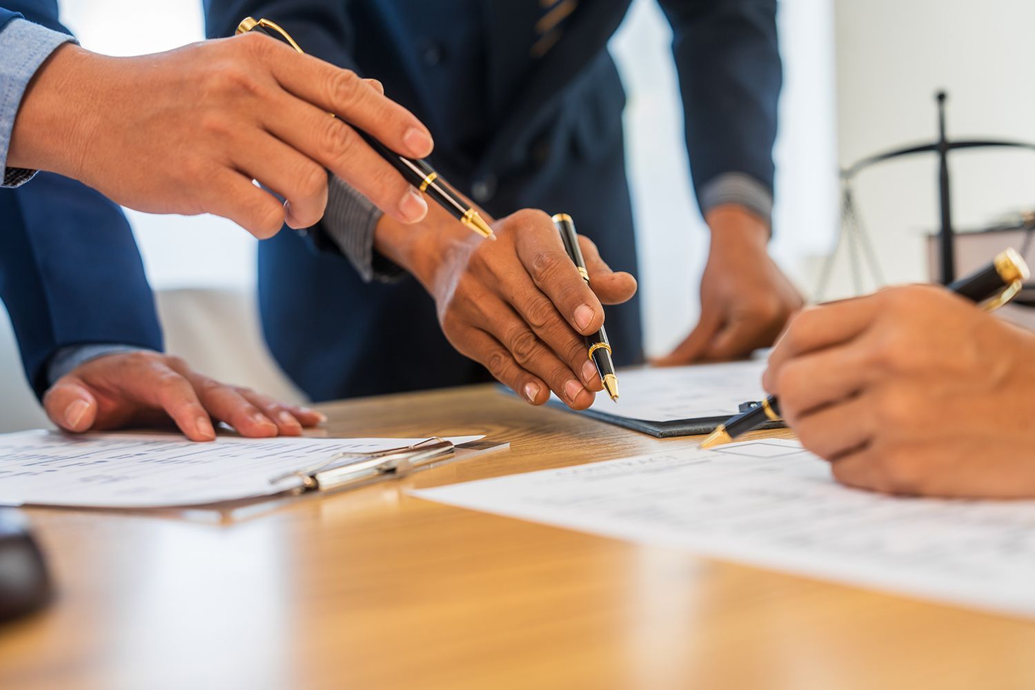 Hands pointing at documents on a wooden table. Three people wearing suits, signing a contract. Hands pointing at documents on a wooden table. Three people wearing suits, signing a contract.