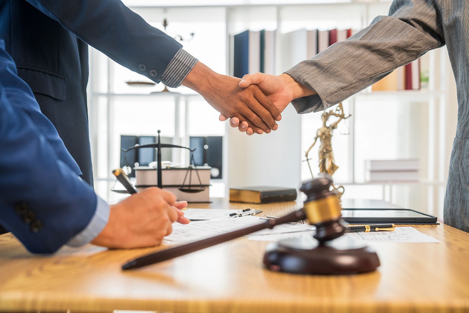 People shaking hands over a desk with a gavel, scales of justice, and documents. People shaking hands over a desk with a gavel, scales of justice, and documents.