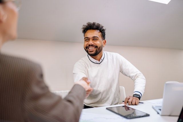 person in brown blazer and another in white sweater shaking hands across a table