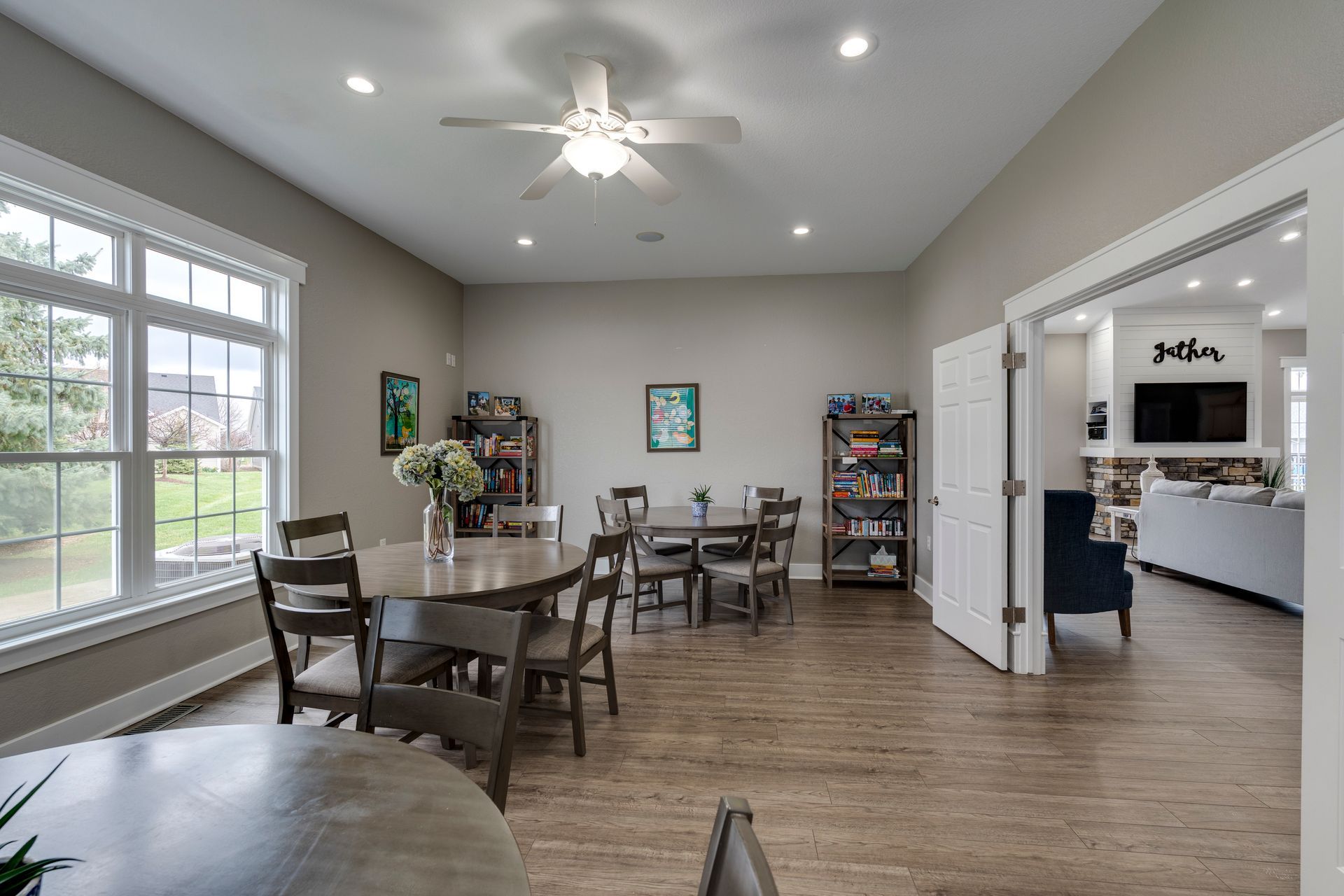 Spacious dining area with round tables, chairs, bookshelves, and large windows. View into a living room with a fireplace.