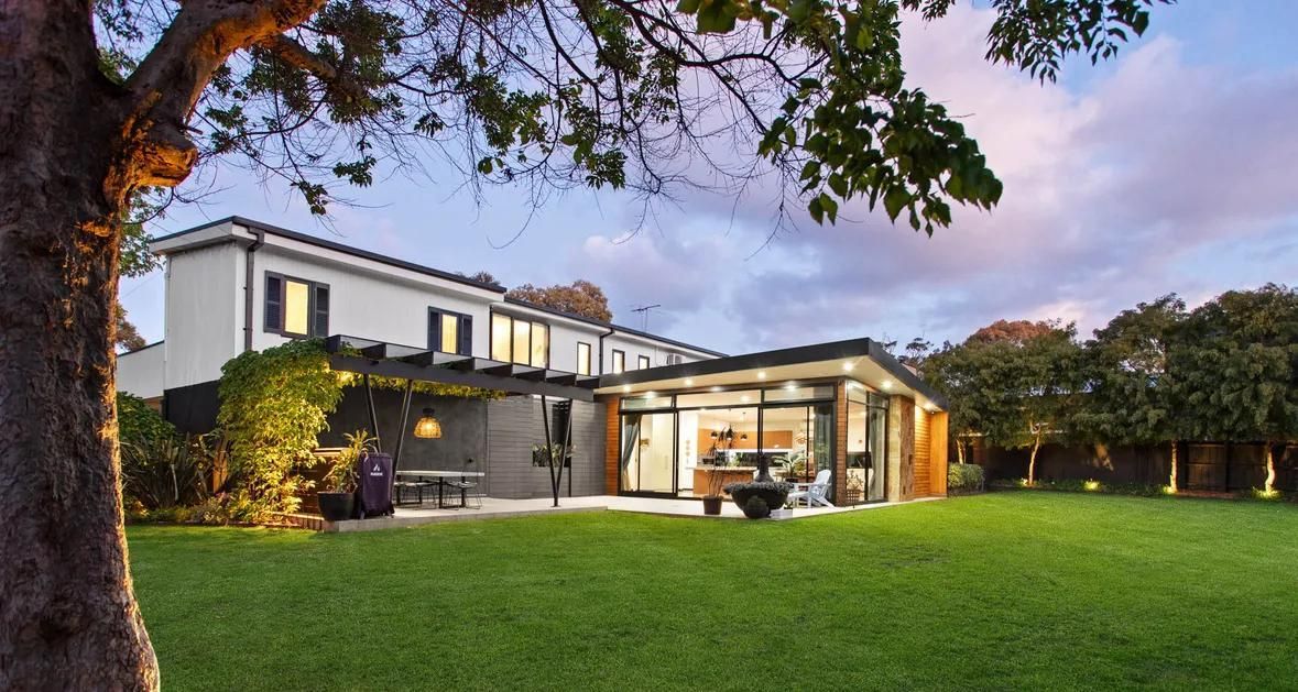 Modern two-story house with a glass-walled patio and lit backyard lawn at dusk