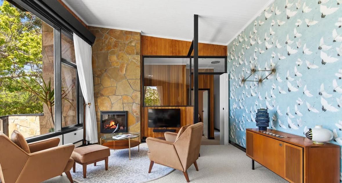 Modern living room with stone fireplace, large windows, blue fish-patterned wall, and tan chairs