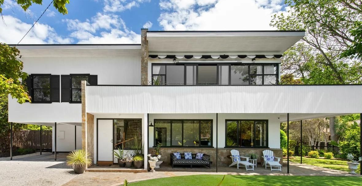 Modern white two-story house with large windows, patio seating, and a green lawn
