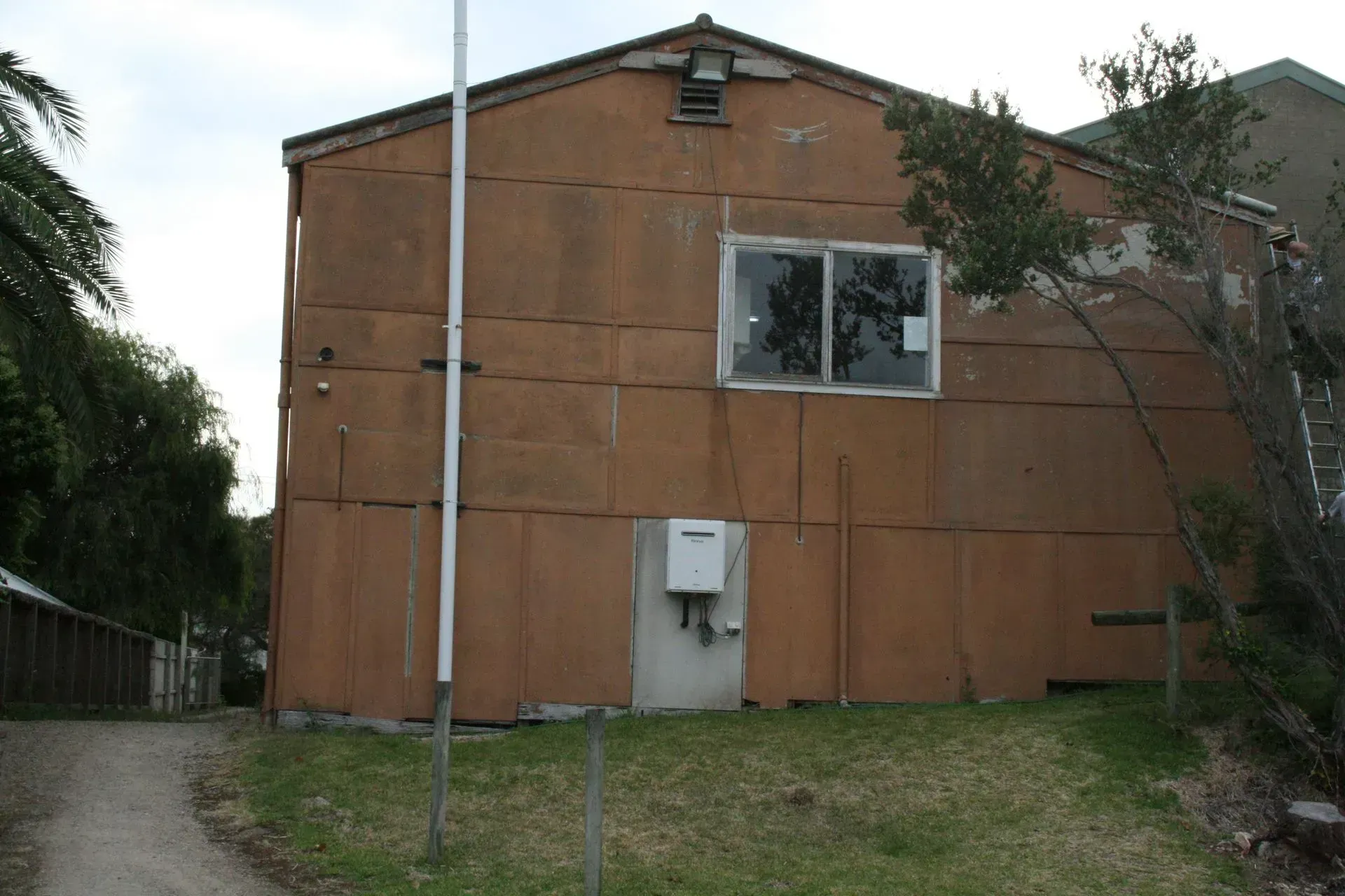 Orange-brown building with a small window and door, seen in a grassy yard beside a pole and trees.