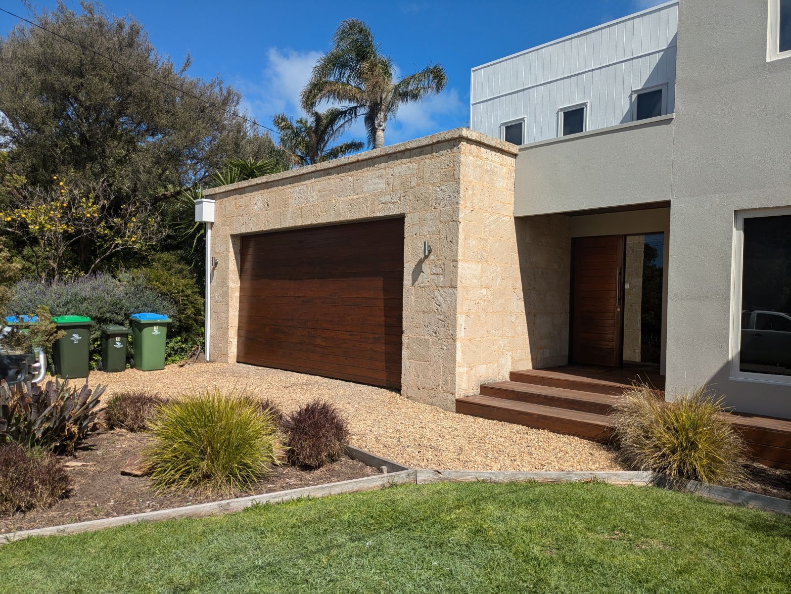 Modern house exterior with stone garage, steps, garden beds, and a green lawn.