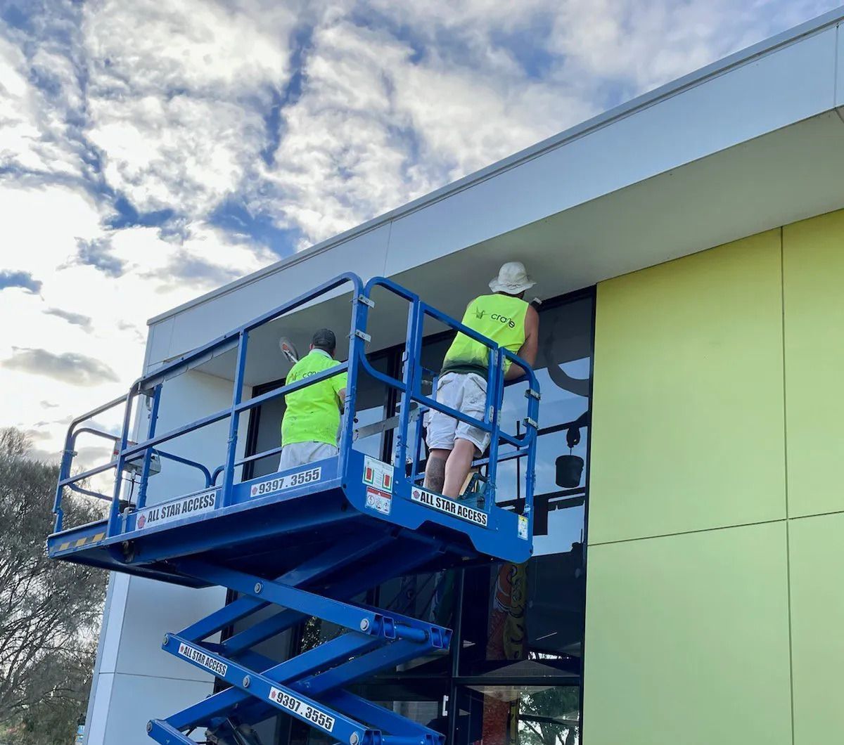 Two workers on a blue scissor lift paint a building’s exterior wall beside a window.