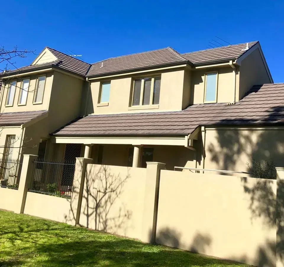Beige two-story townhouse with tiled roof, windows, and a grassy yard behind a fence in bright sunlight
