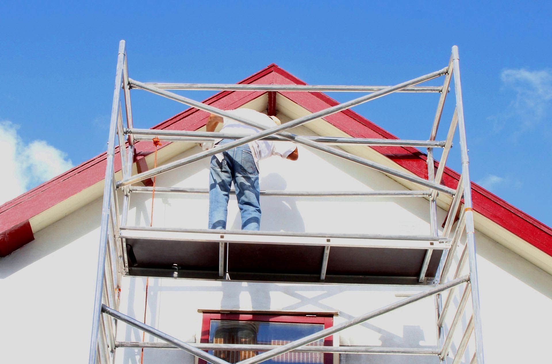 Worker on scaffolding painting a white house with a red roof against a blue sky