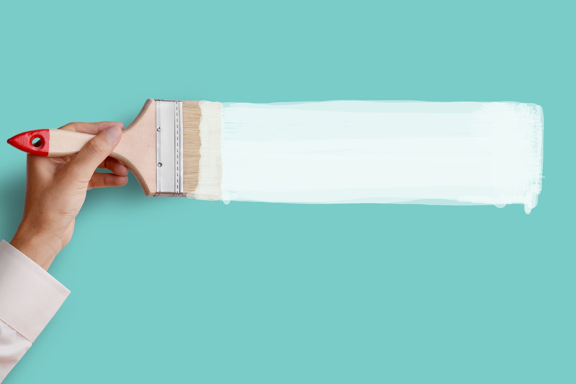 Hand holding a paintbrush applying white paint on a teal background