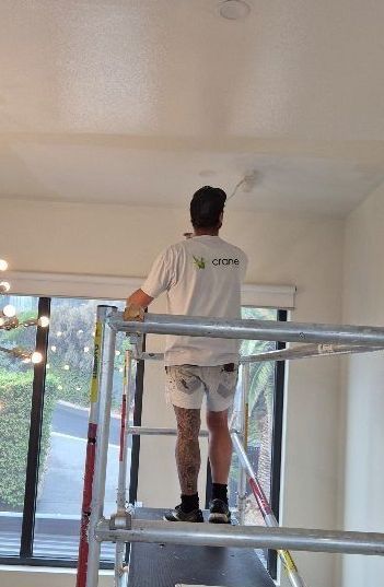 Person painting a white ceiling while standing on scaffolding indoors near a large window.