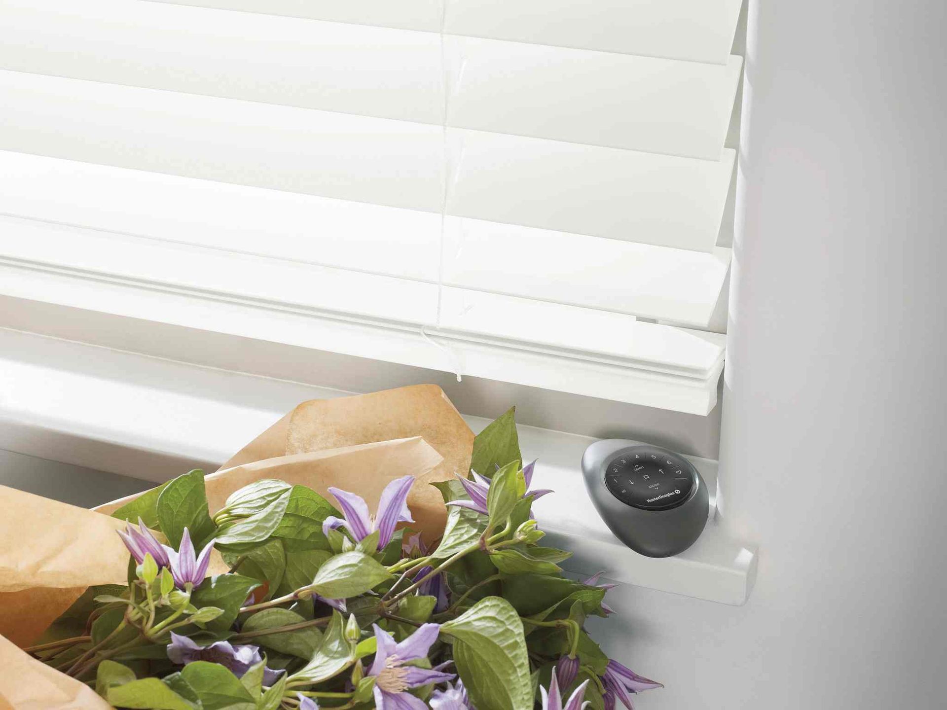 Black Hunter Douglas PowerView® Pebble® Remote on a window sill beneath faux wood blinds and flowers at Alton Bay Blinds near Hampton, NH