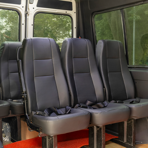 Interior de uma van com quatro assentos pretos e cintos de segurança. Tapete vermelho no chão.