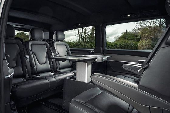 Interior de uma van de luxo com assentos de couro preto, uma mesa e grandes janelas com vista para o exterior.