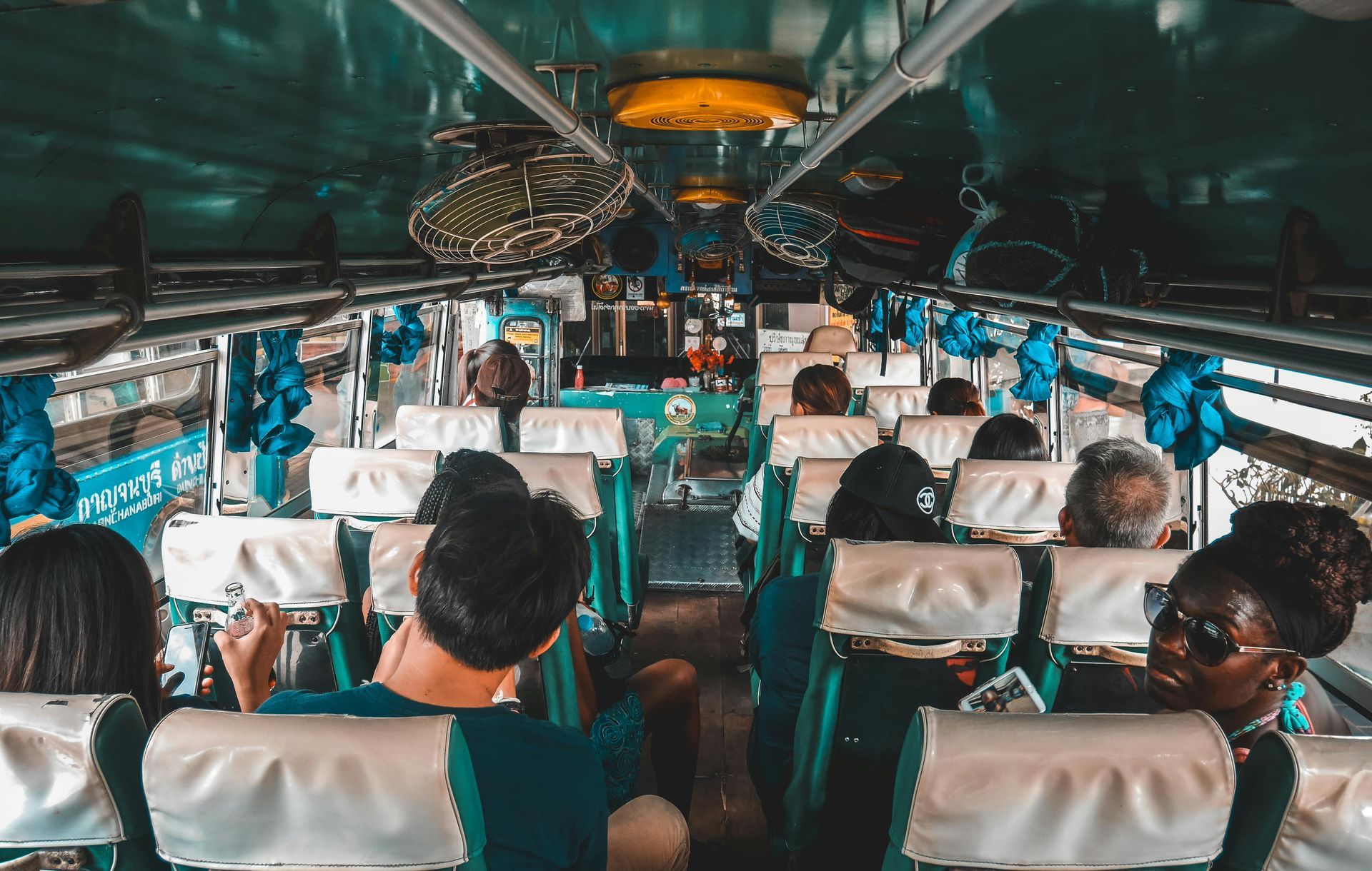 Interior de um ônibus cheio de passageiros sentados, com detalhes em turquesa e azul. Iluminação forte no teto.