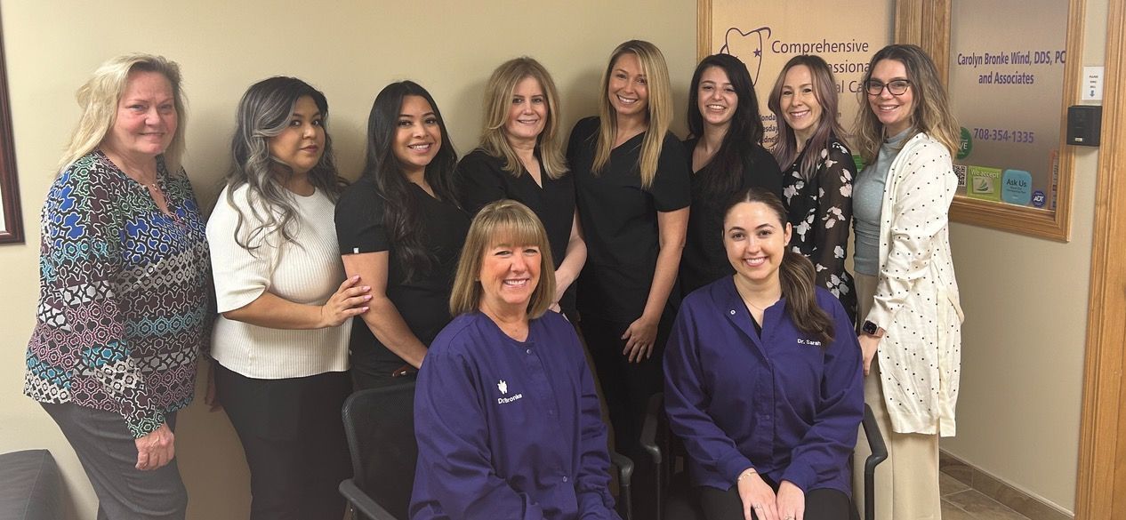Group of smiling women posing indoors. Some wear black scrubs, some blue scrubs. Wall art in background.