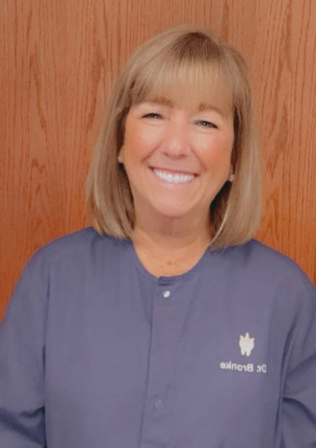 Woman with blonde hair smiling, wearing blue scrubs, likely in a dental office.