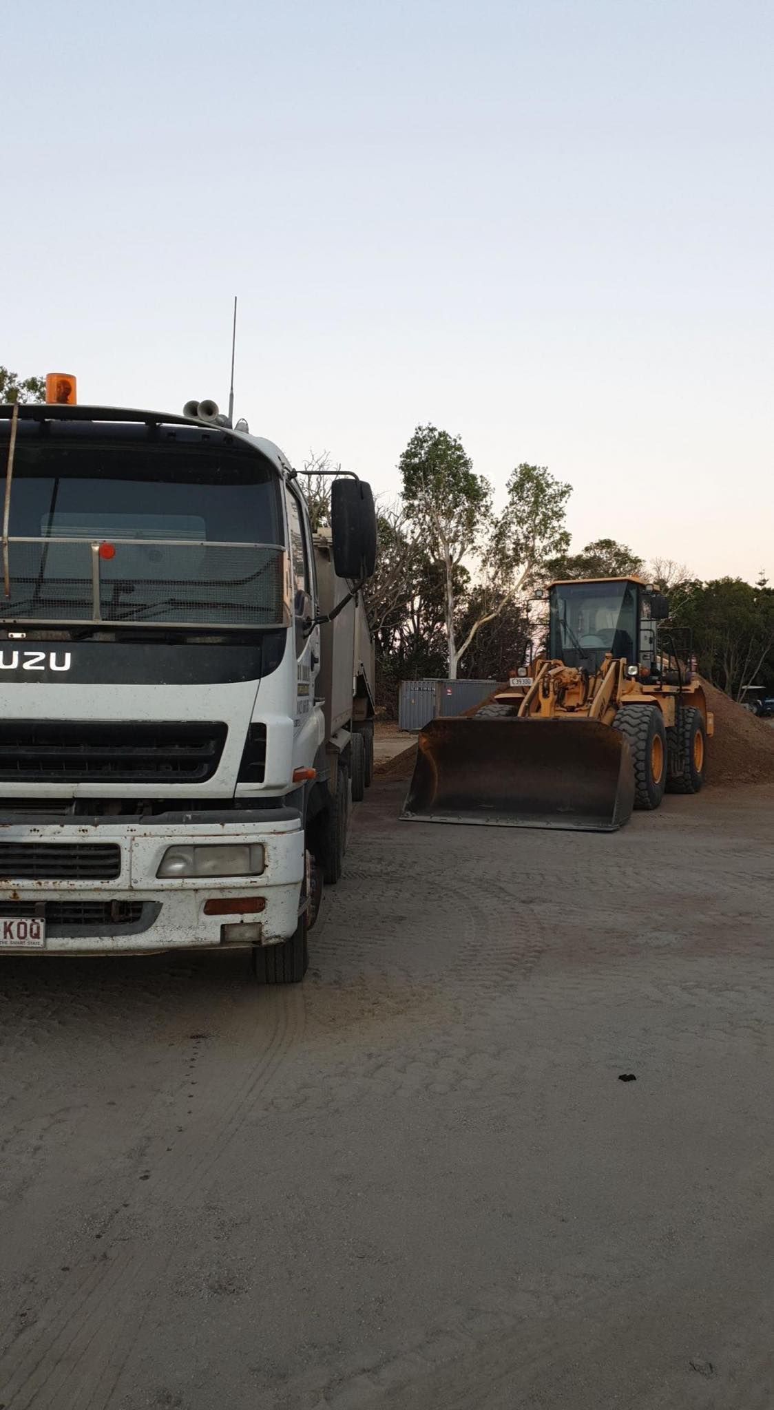 White Isuzu truck next to a yellow front-end loader on a dirt lot; trees and sky in the background. . - DJ Brazil earthmoving, truck and tilt tray hire, Bowen QLD