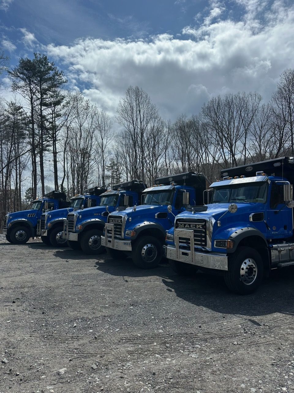 Row of blue utility trucks parked on gravel under a cloudy sky with bare trees in the background