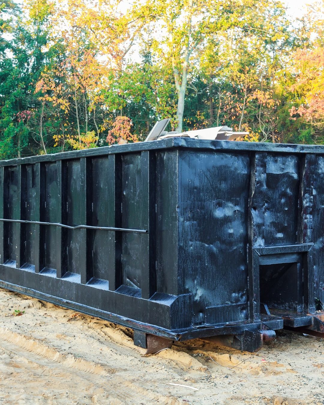 Black dumpster outdoors, near trees.