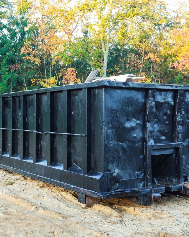 Black dumpster outdoors with fall foliage background.