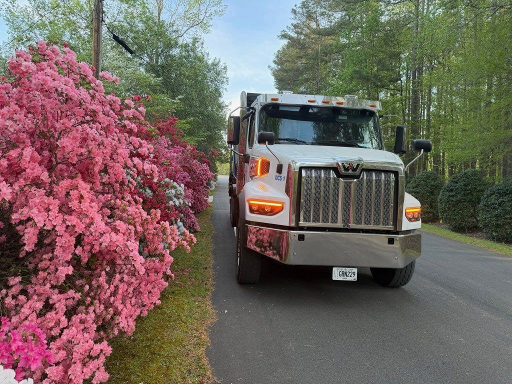 White truck driving on a road beside bright pink flowering bushes and green trees.