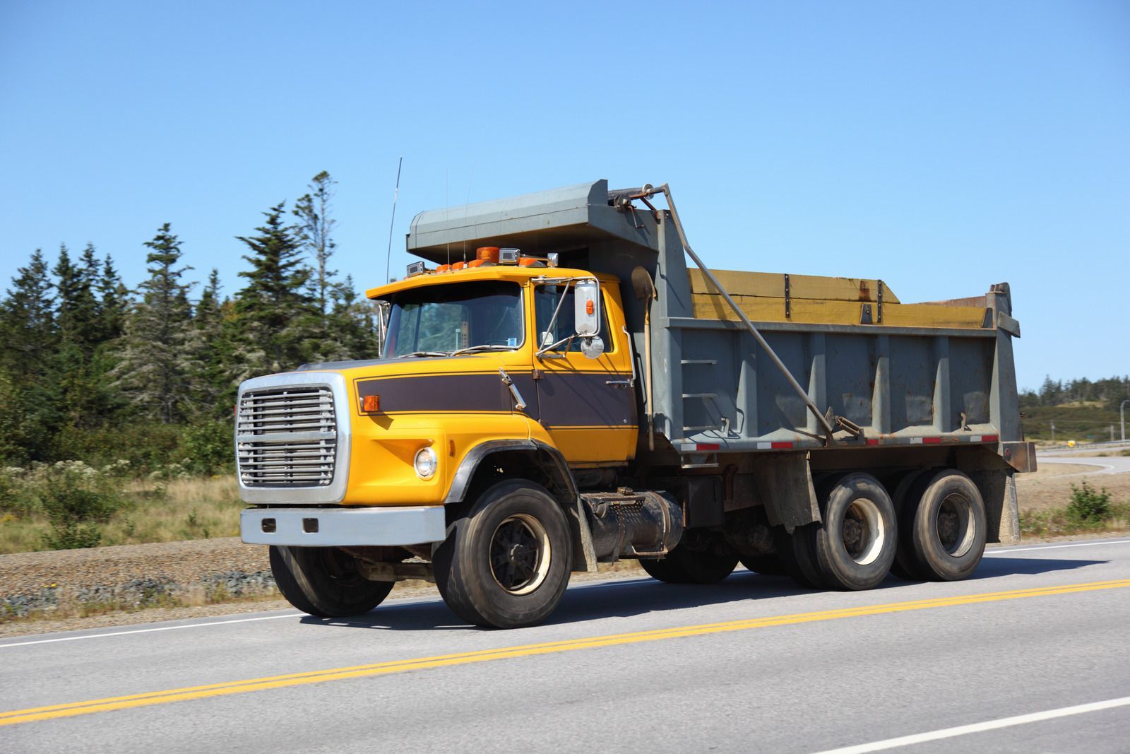 Yellow and gray dump truck driving on a road under a blue sky, hauling wooden planks.