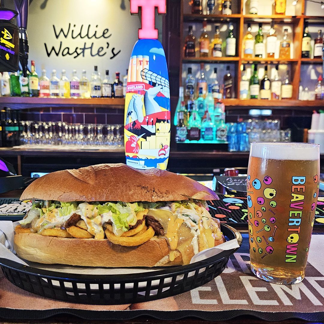Sandwich and beer on a bar table, with colorful bottles and glasses behind them