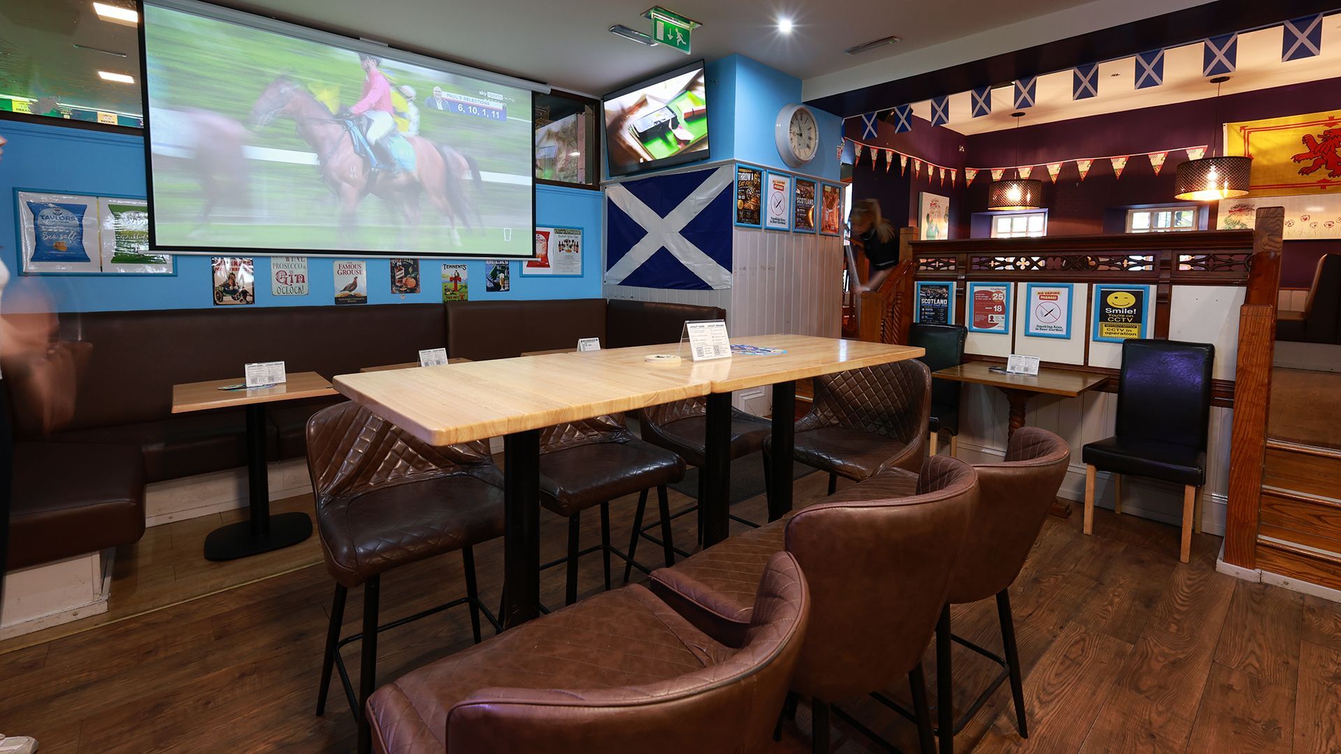 Cozy pub interior with TV screens, high tables, leather chairs, and a large Scottish flag on the wall
