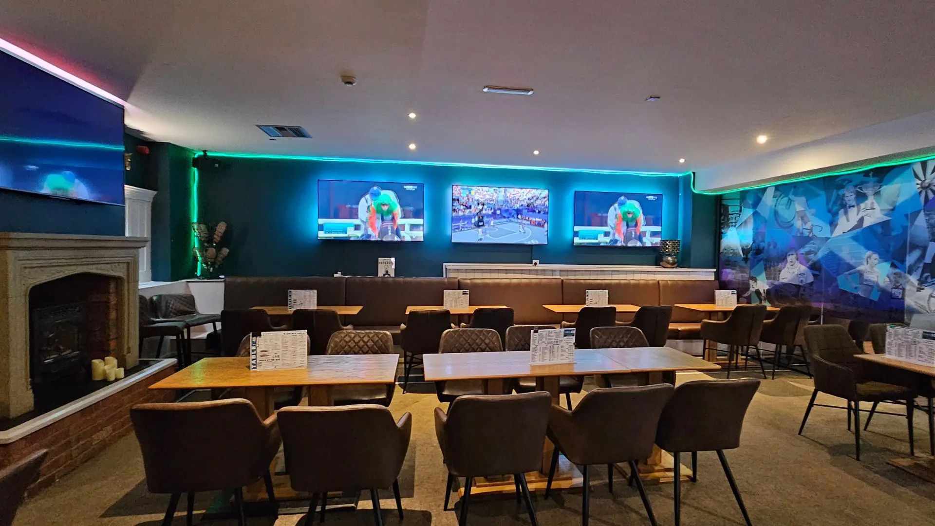 Empty restaurant dining room with tables and chairs, blue-lit bar, fireplace, and large wall screens