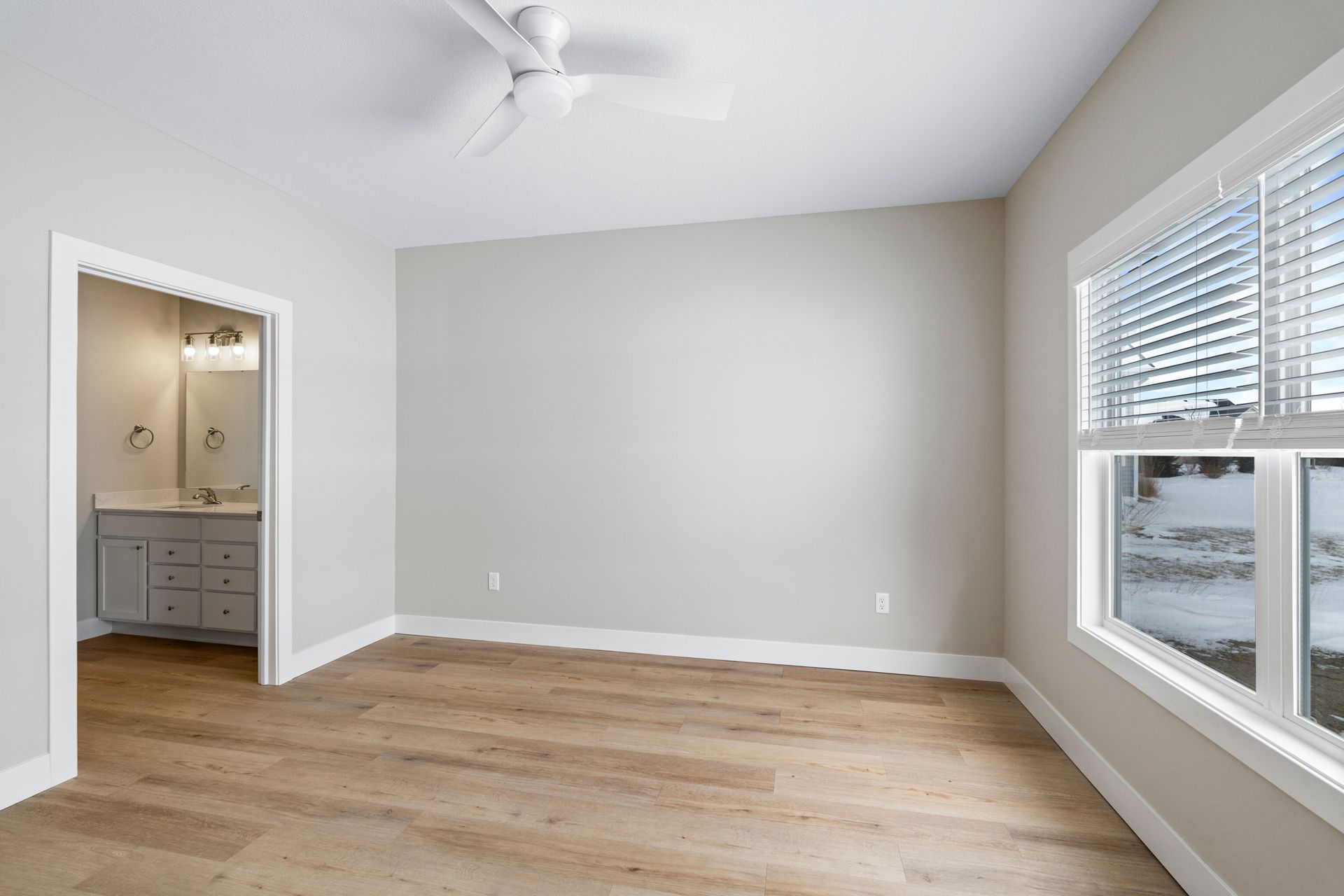 Empty bedroom with light wood floors, neutral walls, and a window; bathroom visible in the doorway.