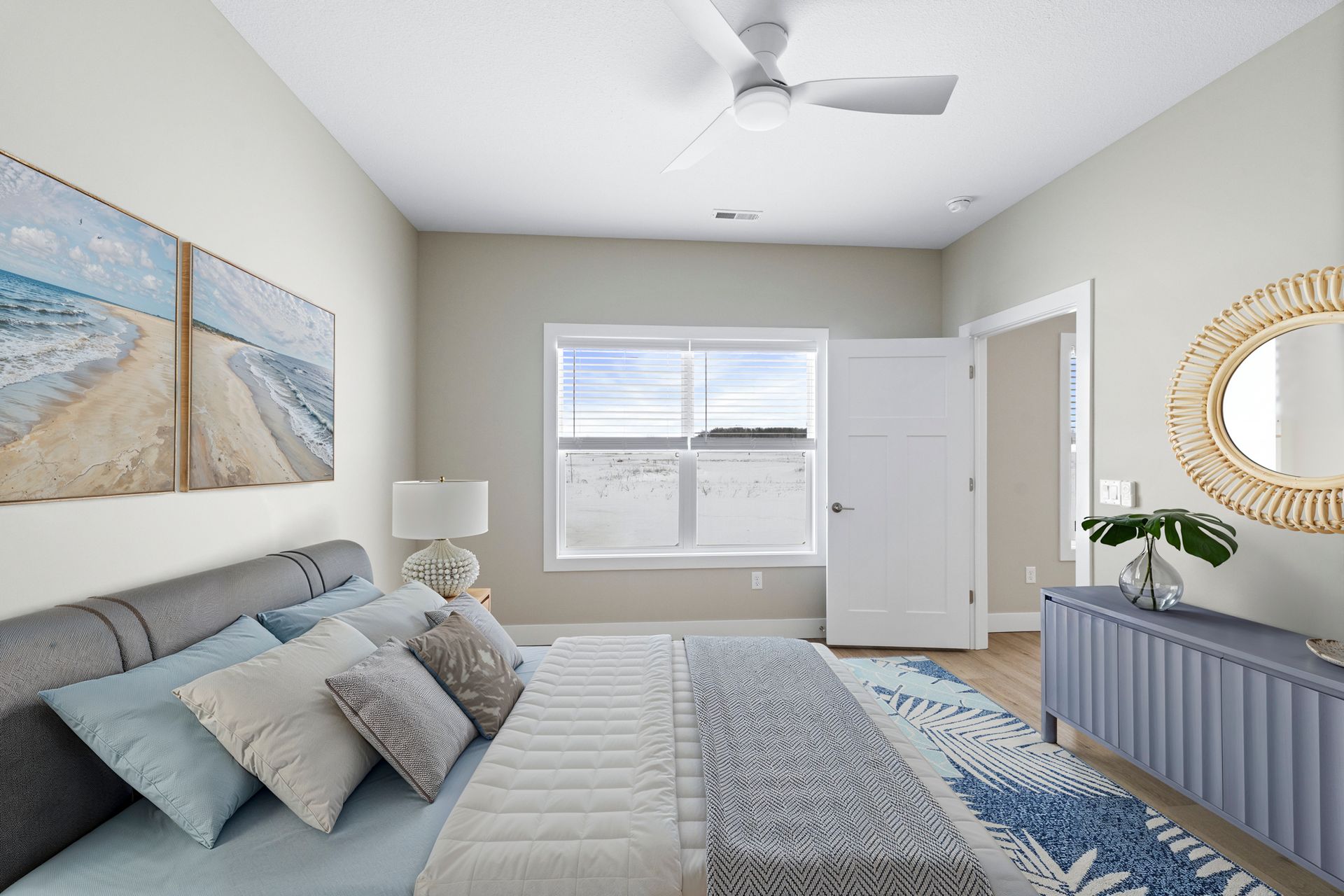 Bedroom with bed, blue dresser, beach art, large window, round mirror, neutral walls.