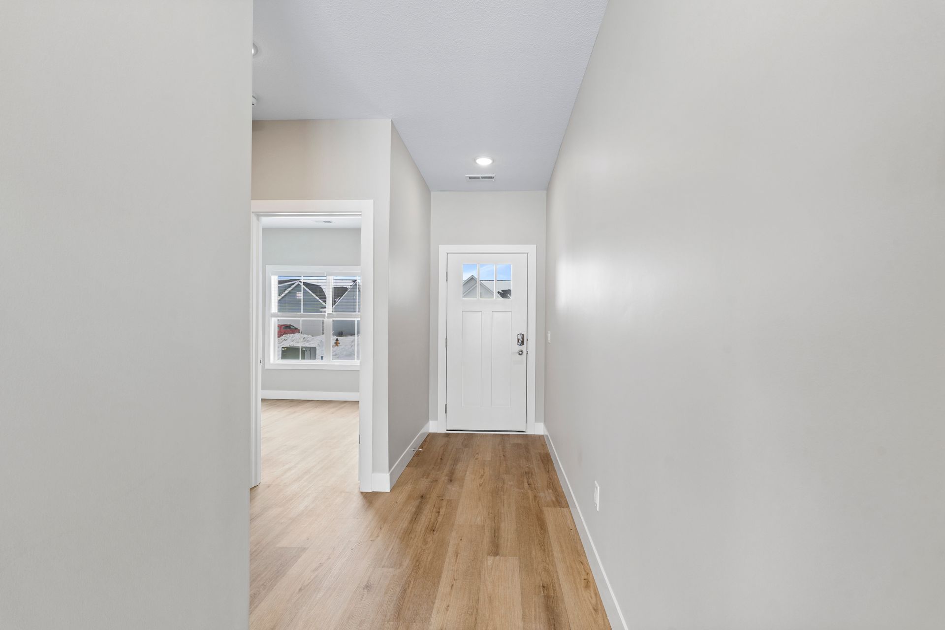 Narrow hallway with light-colored walls and wooden floors. White front door is at the end. Another doorway on the left.