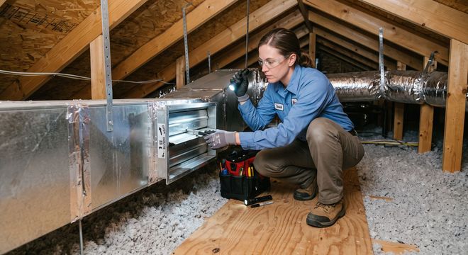 Technician kneeling in an attic, inspecting HVAC ductwork beside a red tool bag.