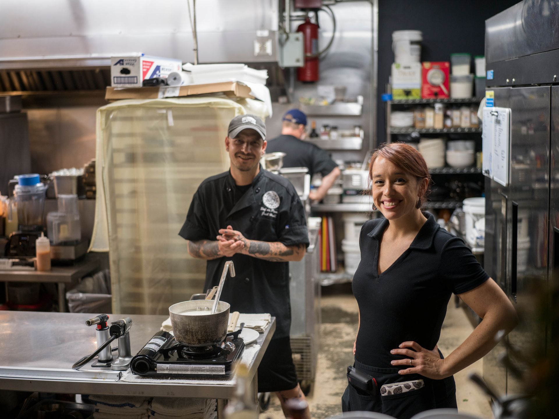 Two people smiling in a commercial kitchen, standing beside a stovetop and cookware.