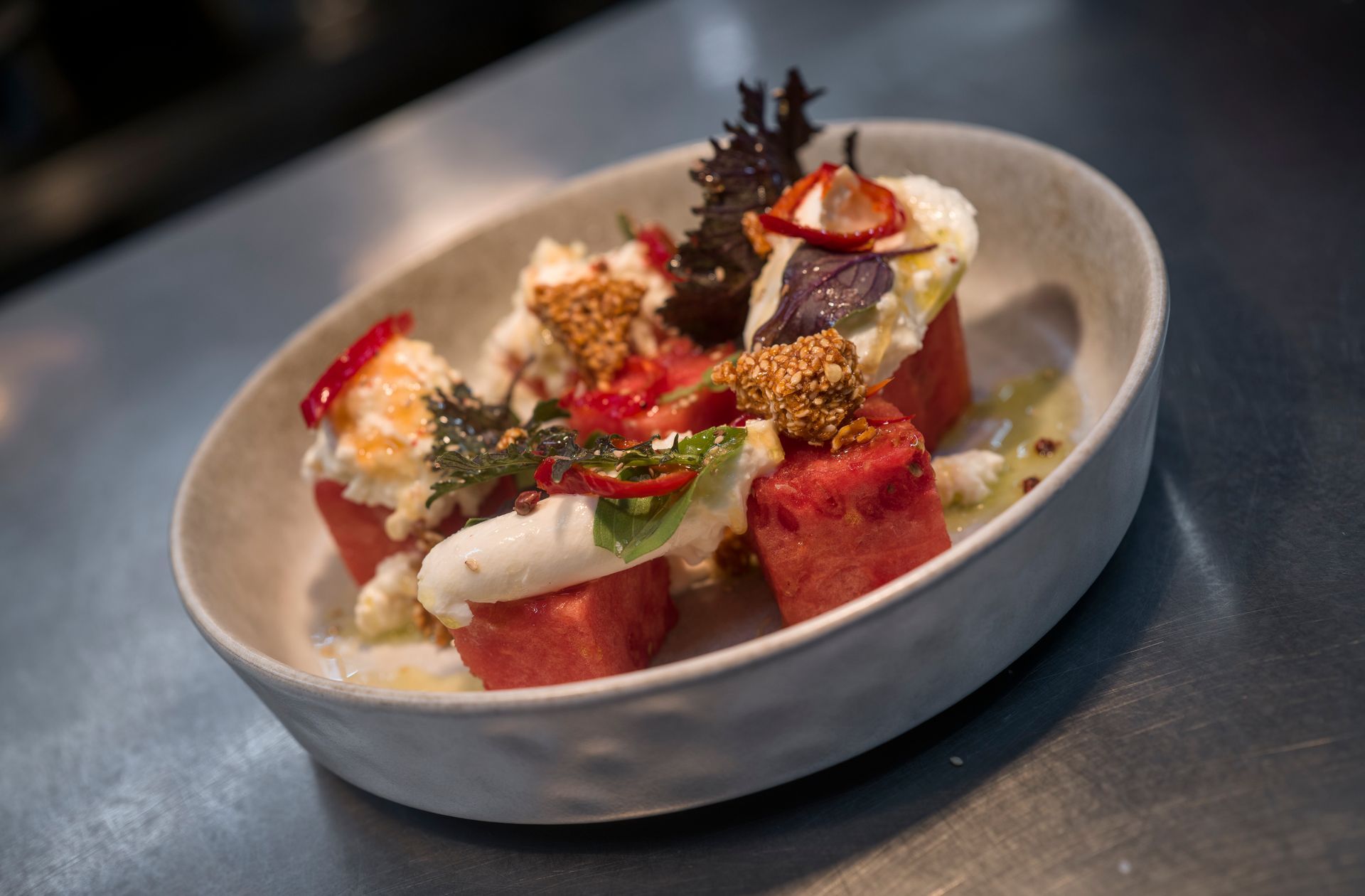 Gourmet plated dessert with watermelon cubes, cream, herbs, and sesame garnish in a white oval dish