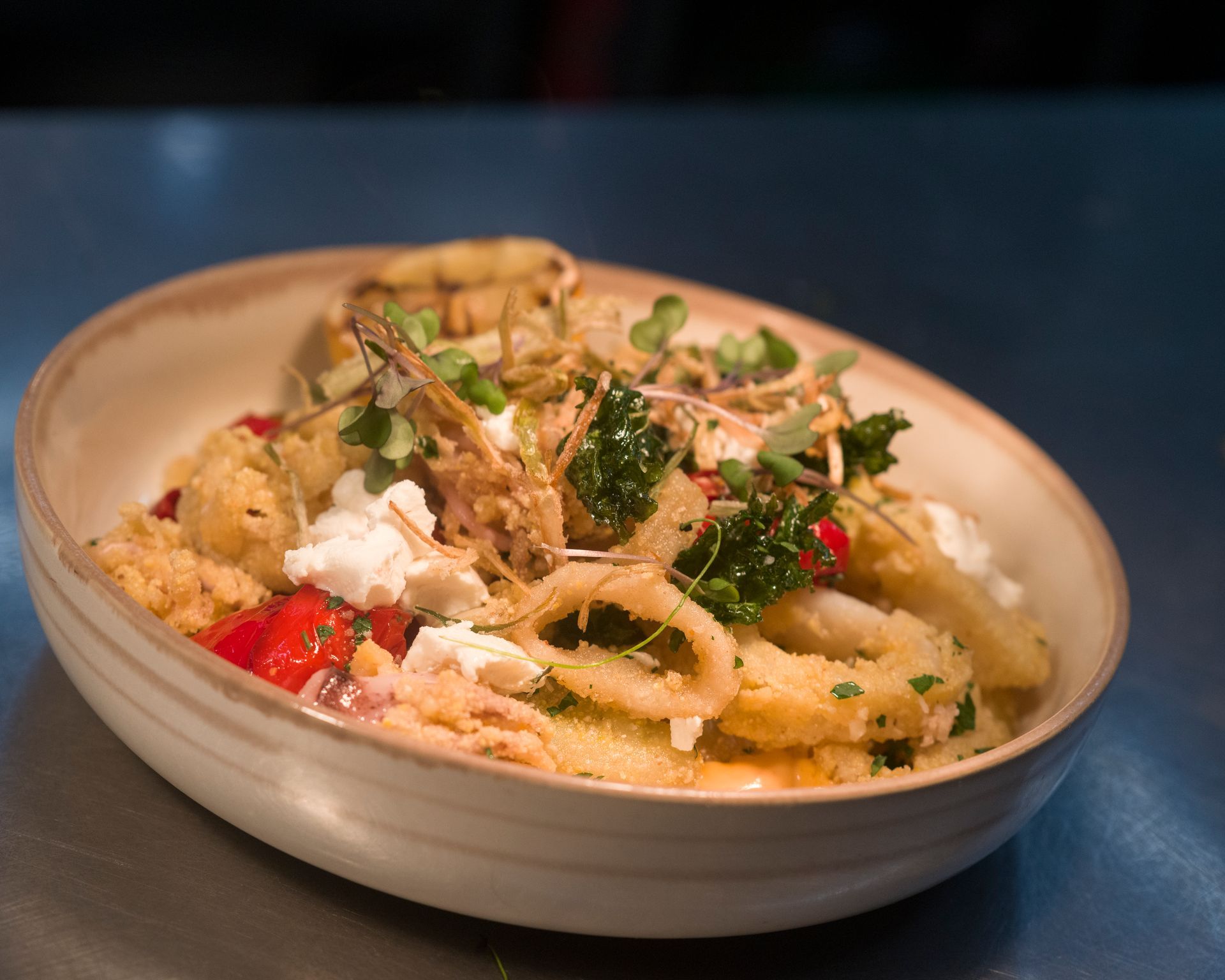 Creamy pasta in a white bowl, topped with herbs, breadcrumbs, and red peppers on a blue table.