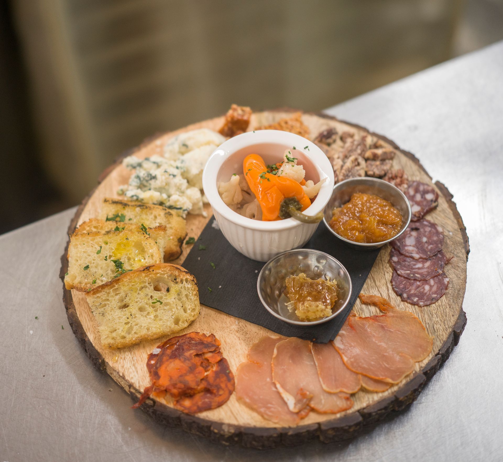 Assorted appetizers on a wooden board with bread, meats, spreads, and a small ramekin of garnish